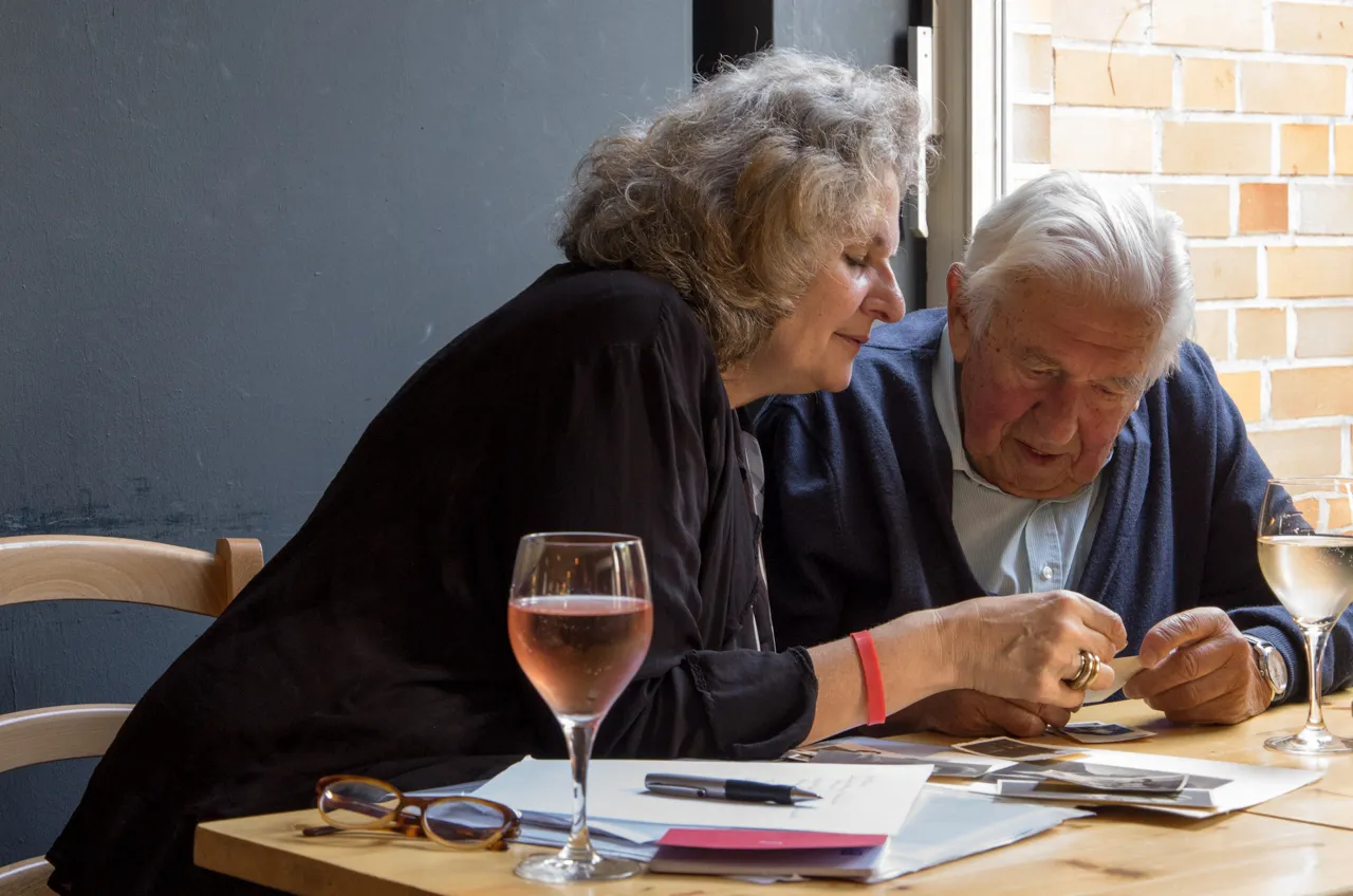 Ulrike Wünnemann und ein älterer Herr sitzen im warmen Fensterlicht zusammen und betrachten eine alte Fotografie.