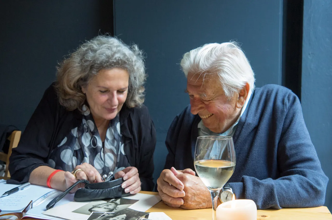 Ulrike Wünnemann im Gespräch mit einem älteren Herrn. Beide sitzen an einem Tisch voller alter Fotografien und lachen gemeinsam.