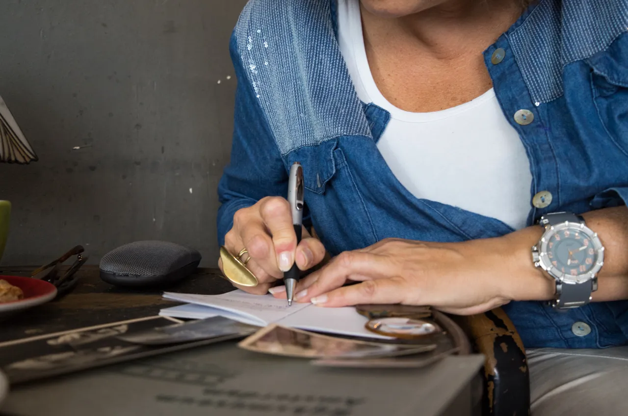 Eine Frau schreibt am Tisch in ein Notizbuch, daneben liegen alte Fotografien.